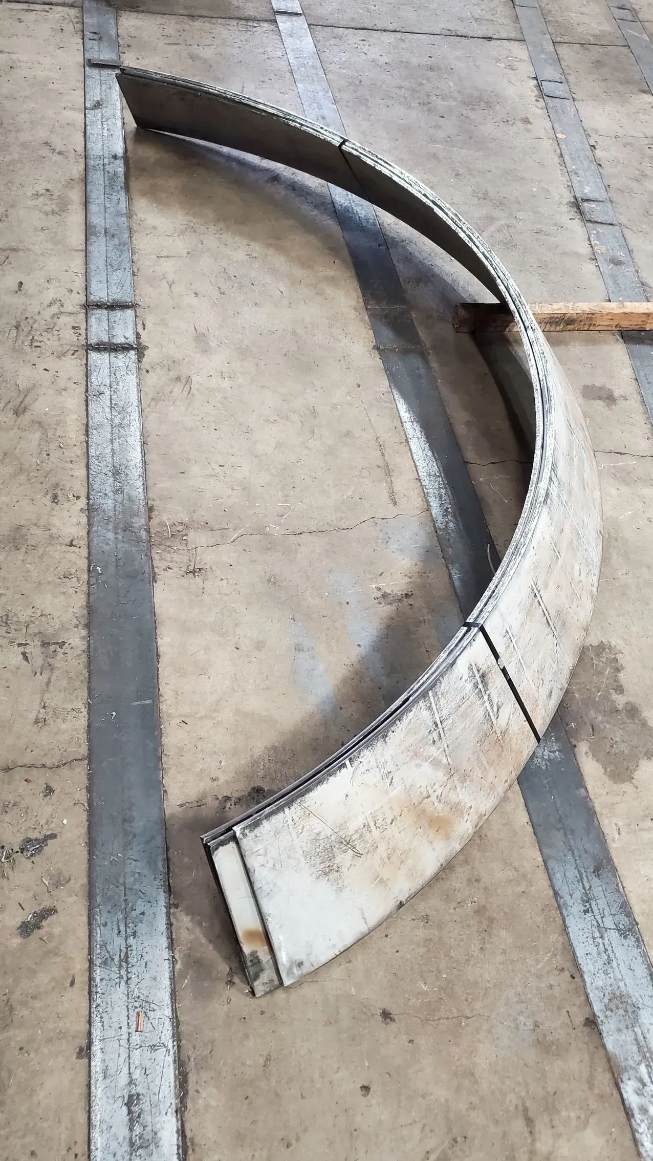 Curved metal component, likely a part of a larger structure, resting on a concrete surface with gray paint.