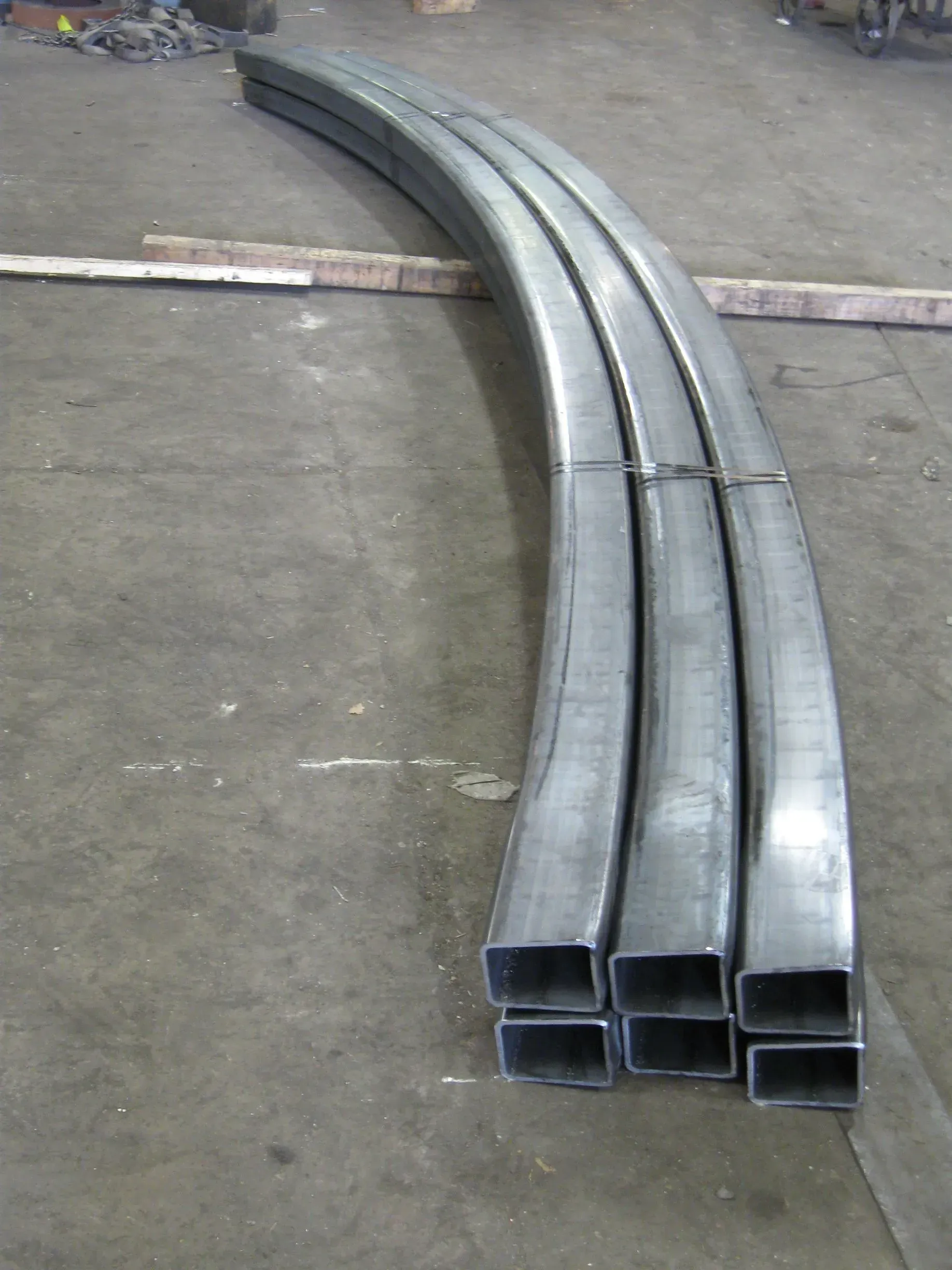 Curved steel tubes, stacked on a concrete floor in a workshop.