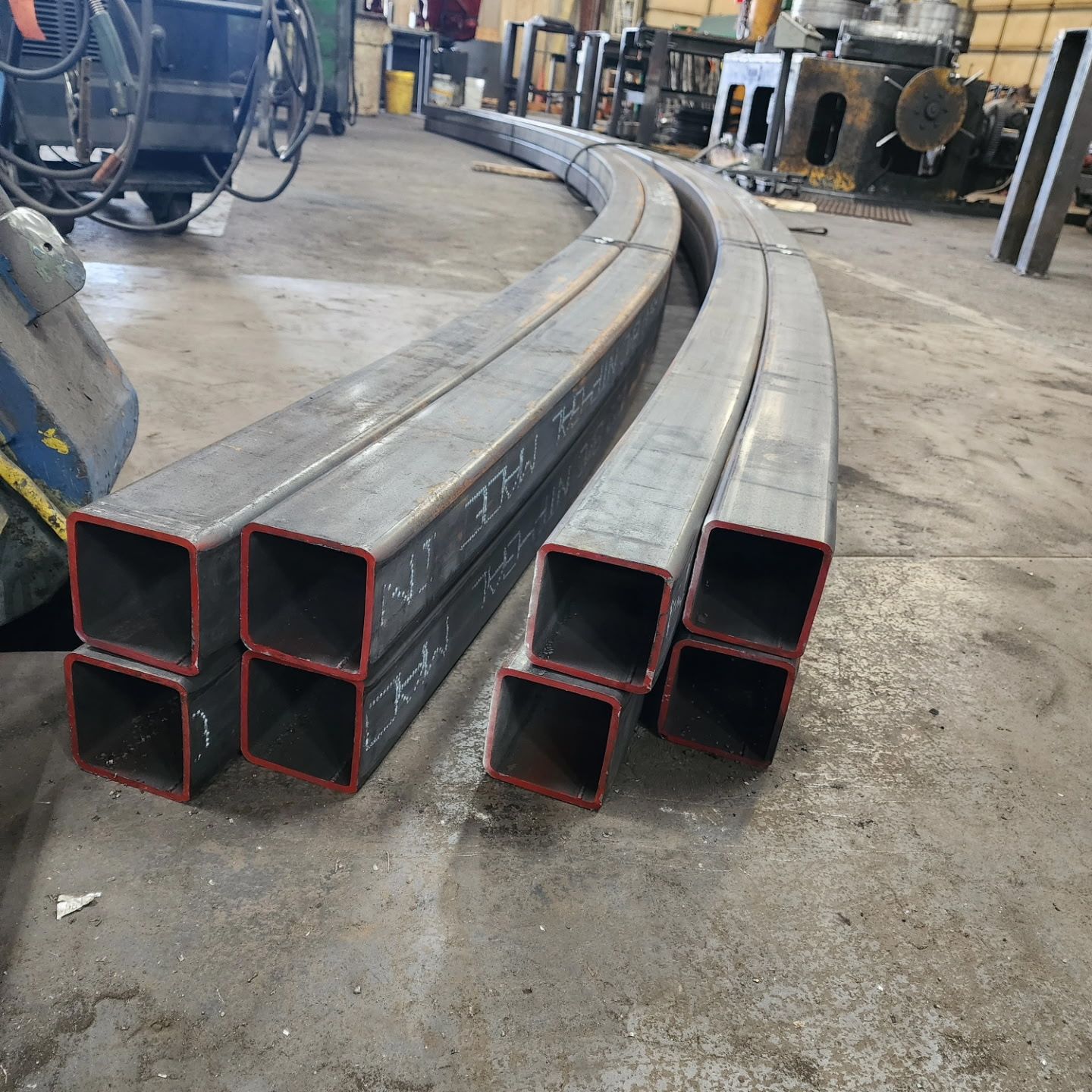Curved, rectangular steel tubes stacked on a concrete floor in a workshop.