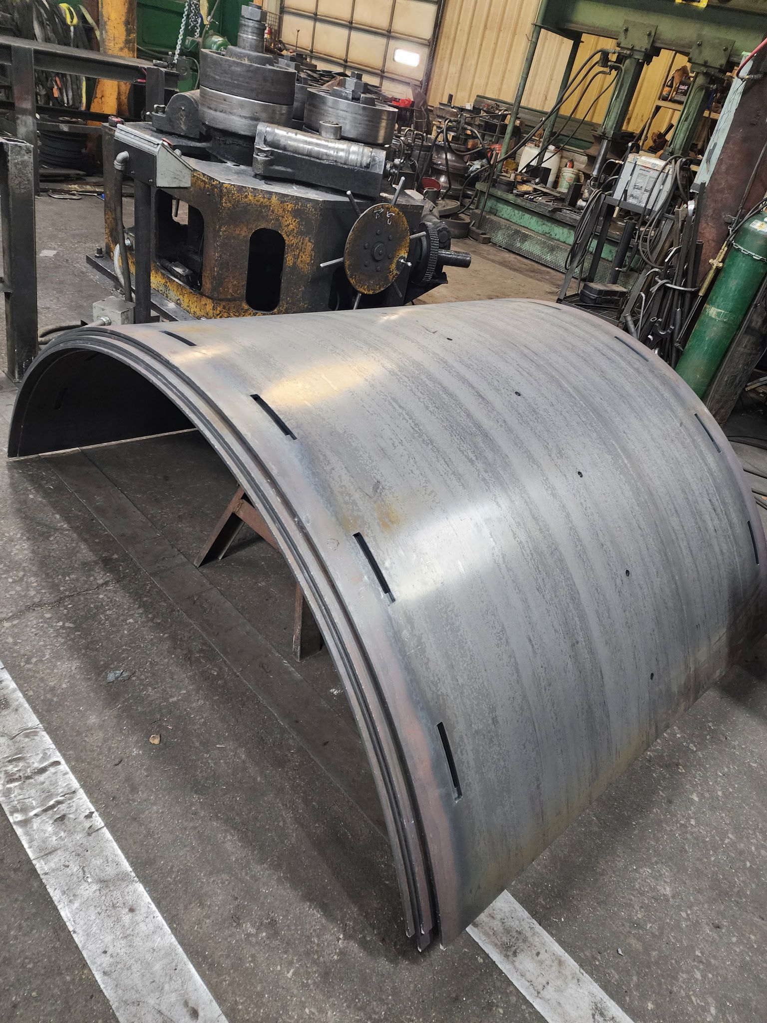 A large, curved metal cylinder in a workshop; it's welded along the edges, and has small cutouts.