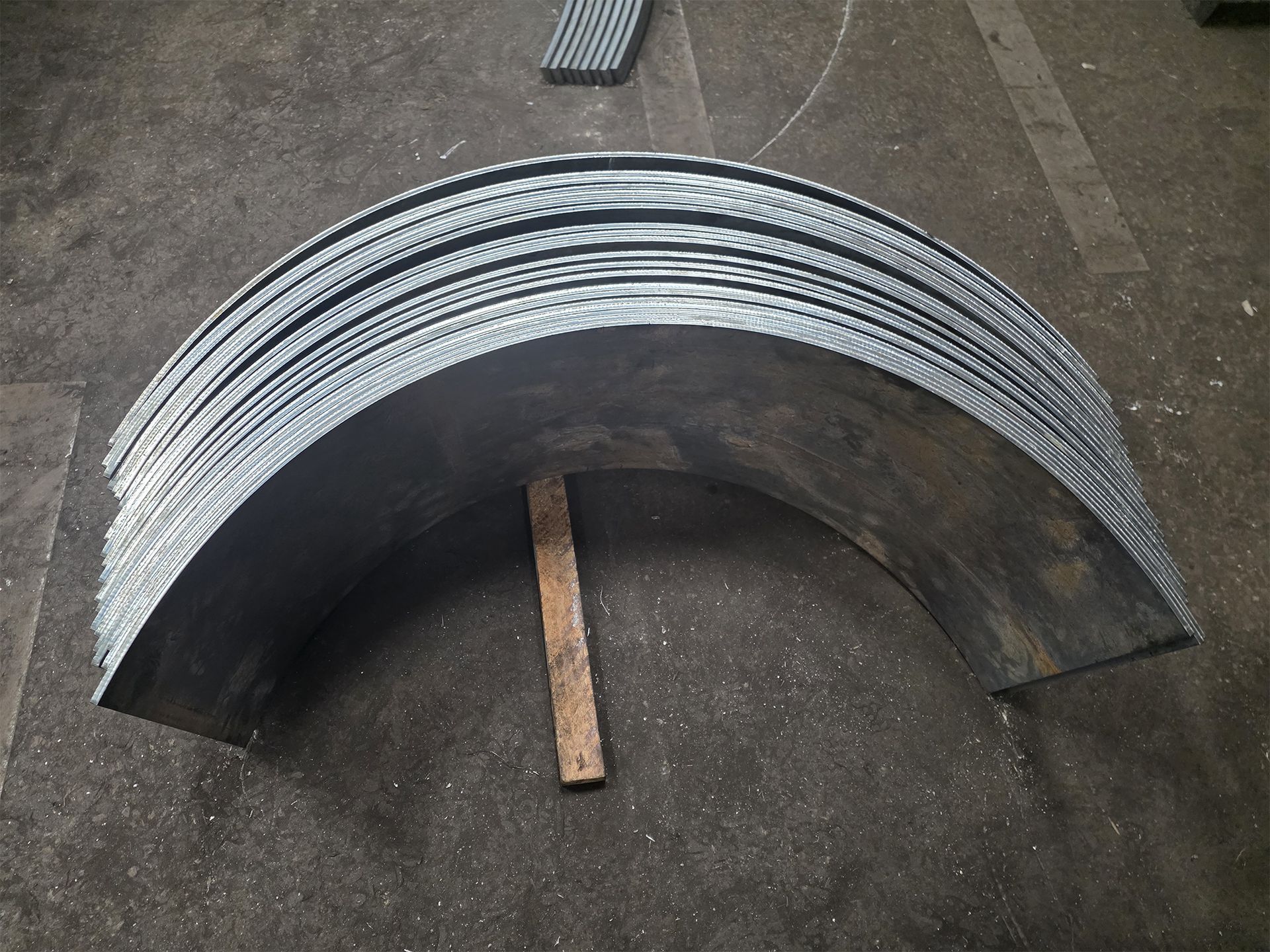 Curved metal sheets stacked, supported by a wooden block on a concrete floor.