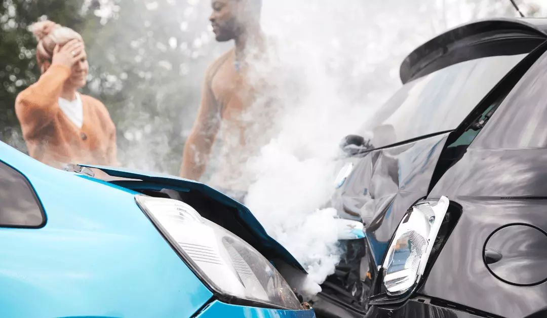 Two cars with crumpled front ends collide on a road, with steam rising between them.