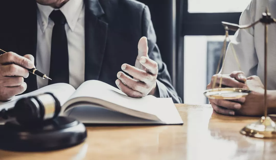 Lawyer speaking at a desk with an open book, gavel, and scales of justice in a courtroom setting