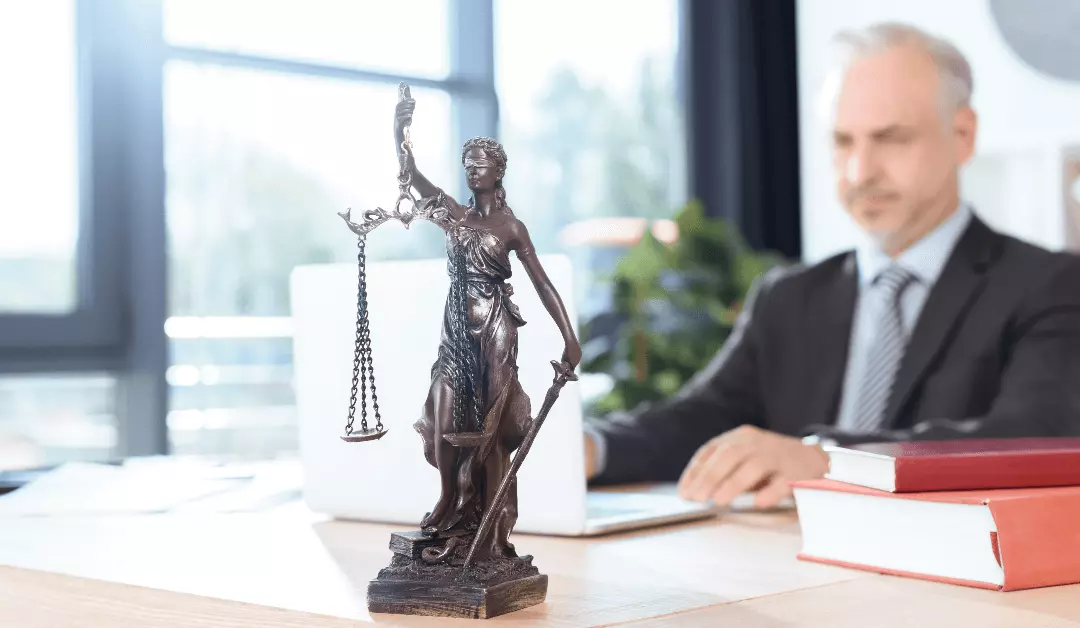 Bronze Lady Justice statue on a desk, with a lawyer blurred in the background and law books nearby