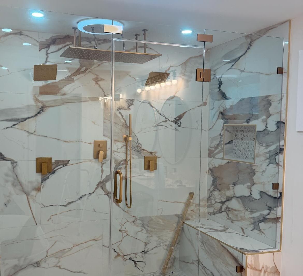 Glass shower enclosure with marble-like wall tiles, gold hardware, and overhead rain shower.