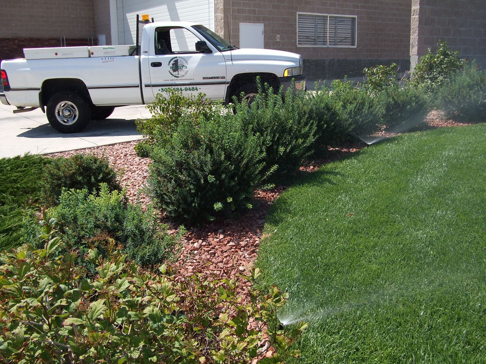 White truck parked near landscaped area with green grass and shrubs; sprinklers running.