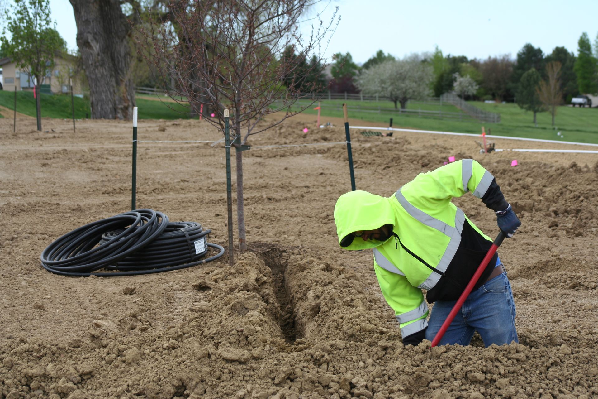 A person wearing a neon yellow jacket digs in soil, near a tree and black hoses.