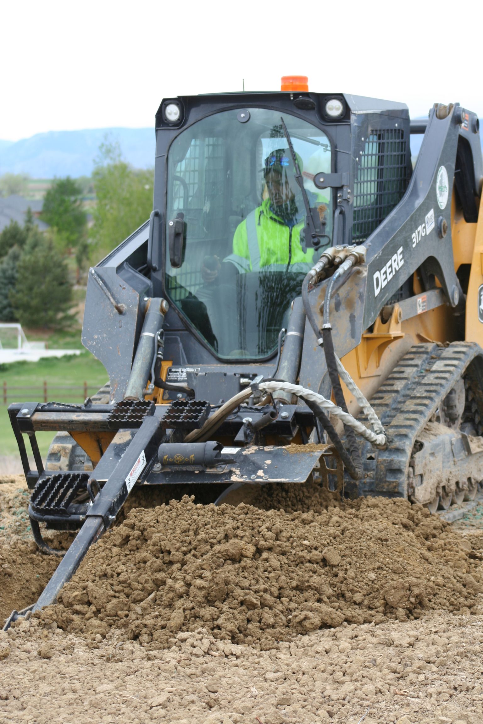 A person in a yellow vest operates a John Deere track loader with a trenching attachment digging into the ground.