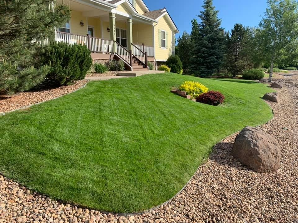 A well-manicured lawn slopes up toward a yellow house. Gravel borders the lawn with large rocks and flowering plants.