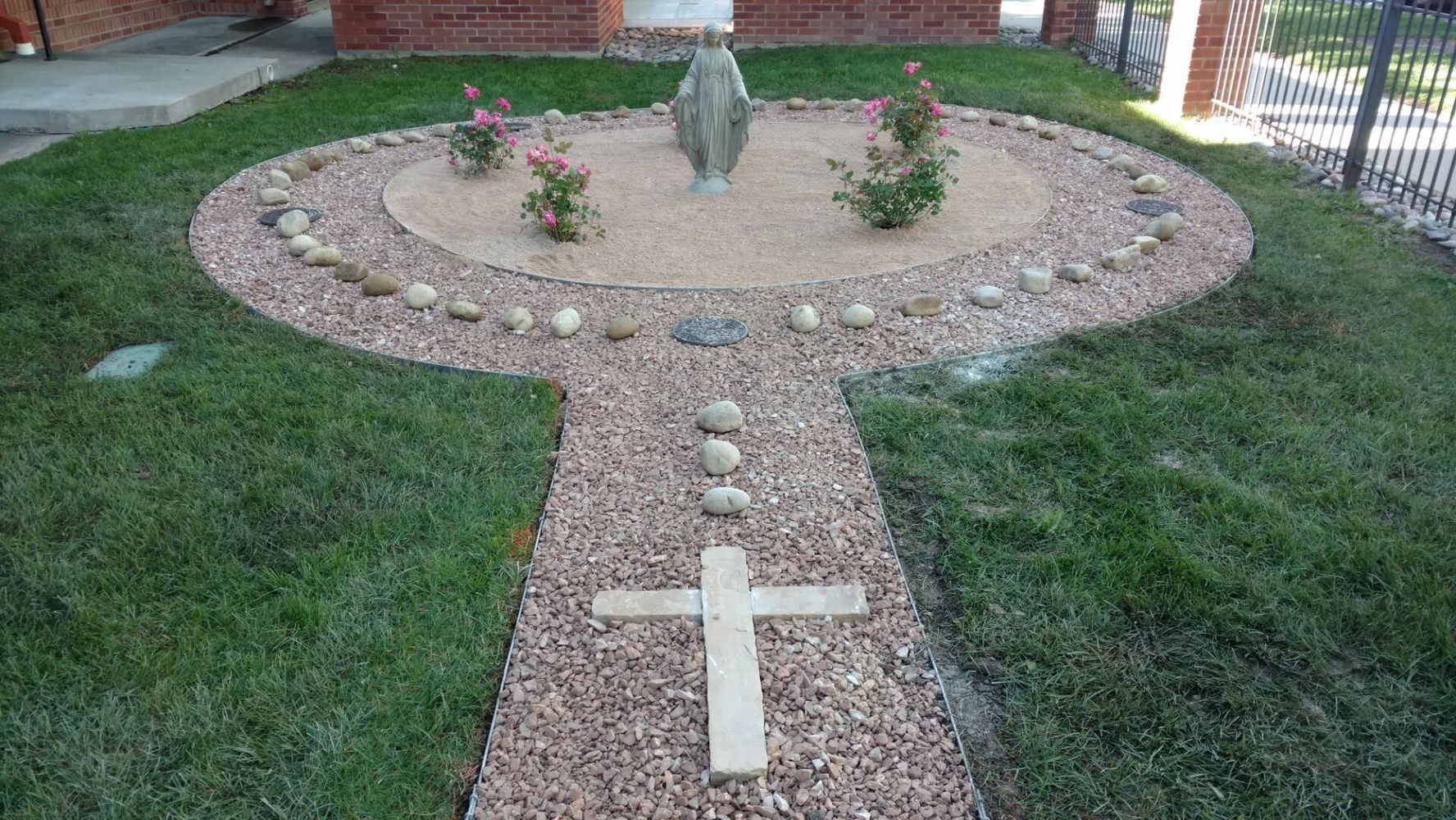 A garden bed in the shape of a rosary with a statue of the Virgin Mary. The pathway to the statue has a cross.