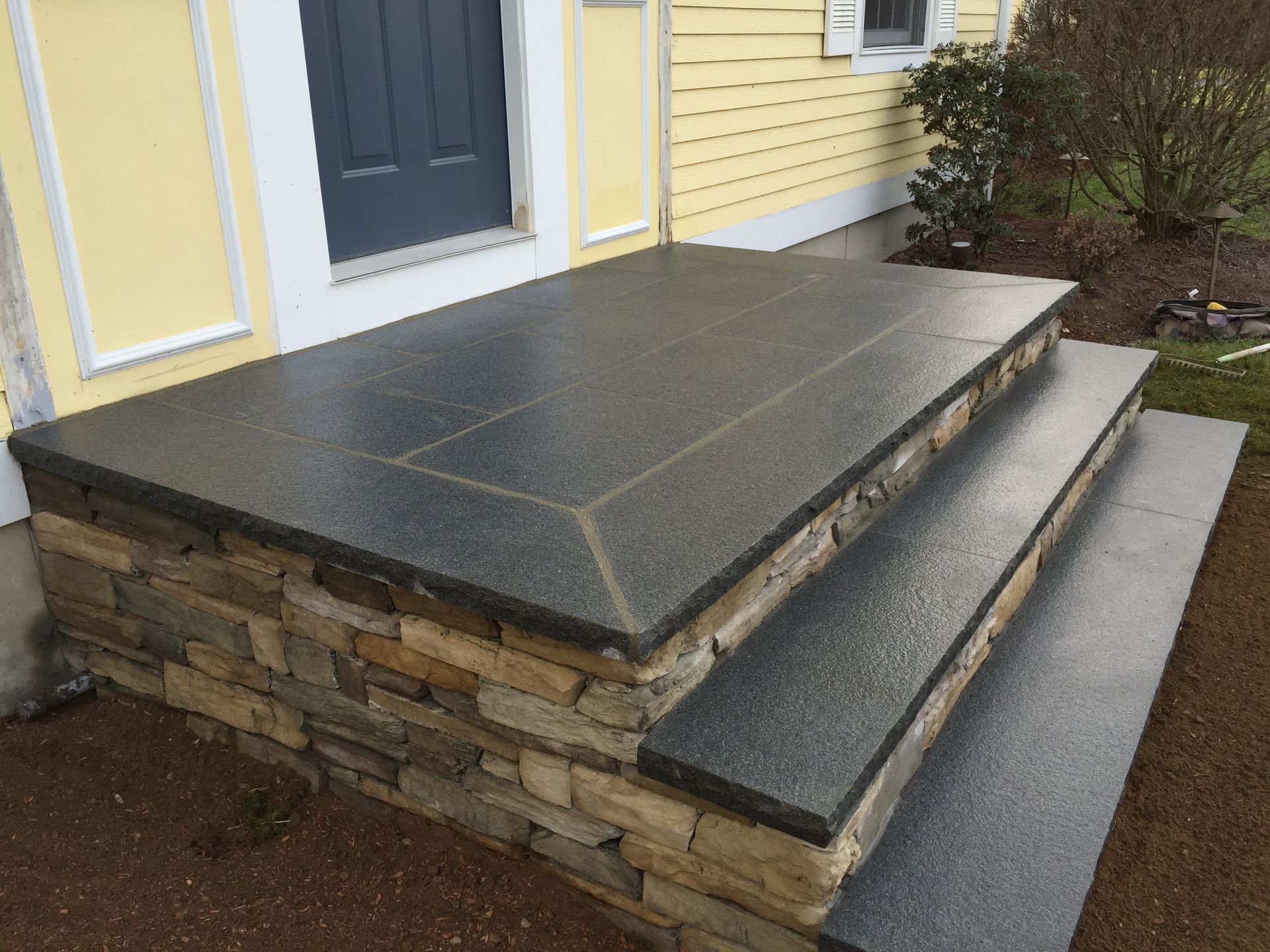 A porch with a stone wall and steps in front of a yellow house.