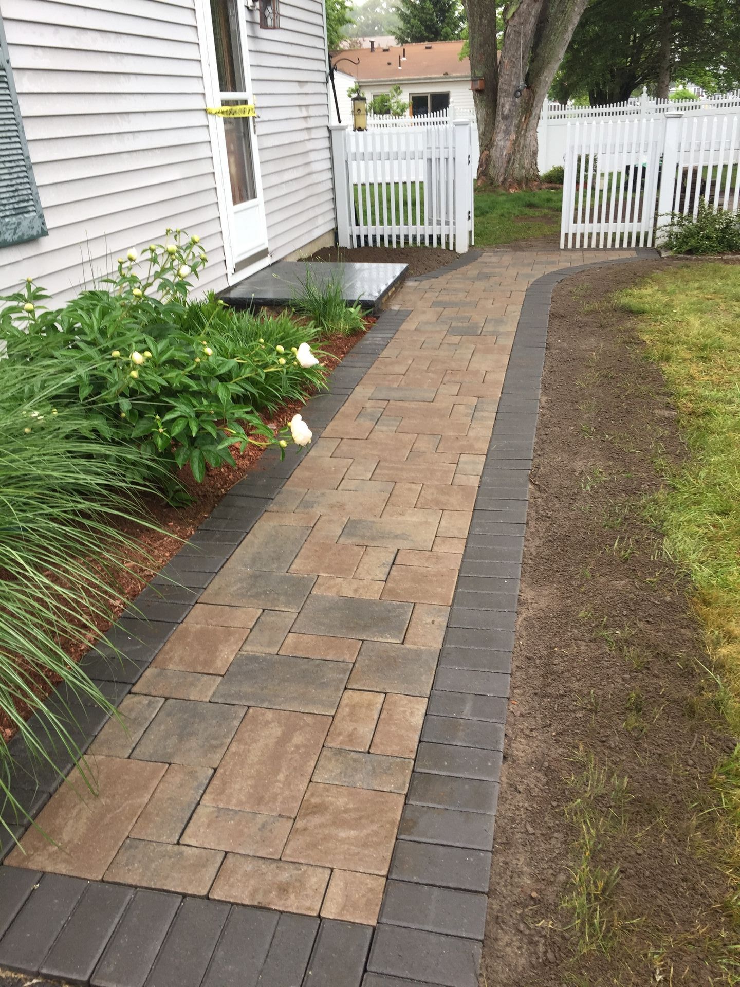 A brick walkway leading to a house with a white fence.