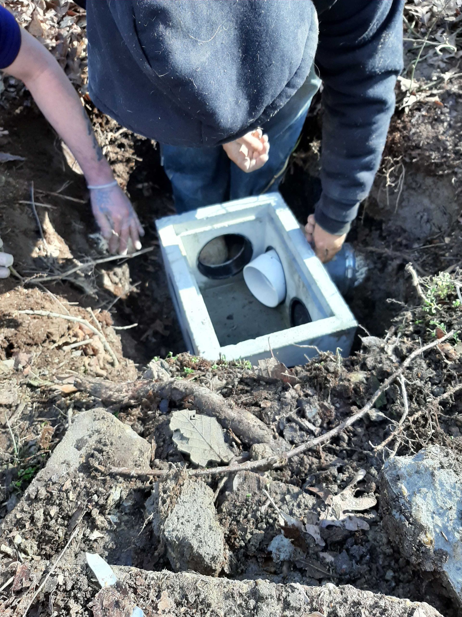 Two people installing a gray box with pipes into a hole in the ground outdoors.