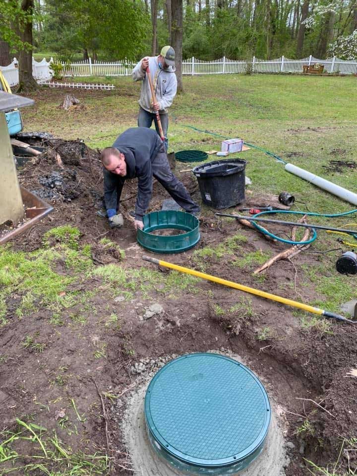 Two people installing a green septic tank cover in a yard; one person is digging, the other is holding a tank ring.