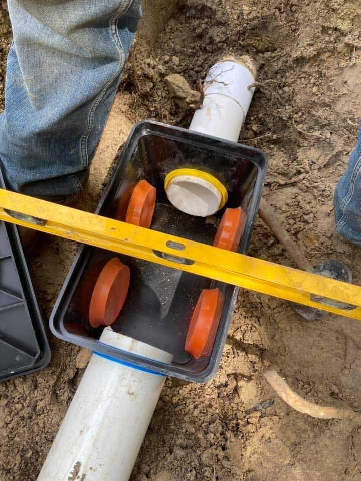 PVC pipe and drain box with orange plugs, being leveled by a person in jeans outside.