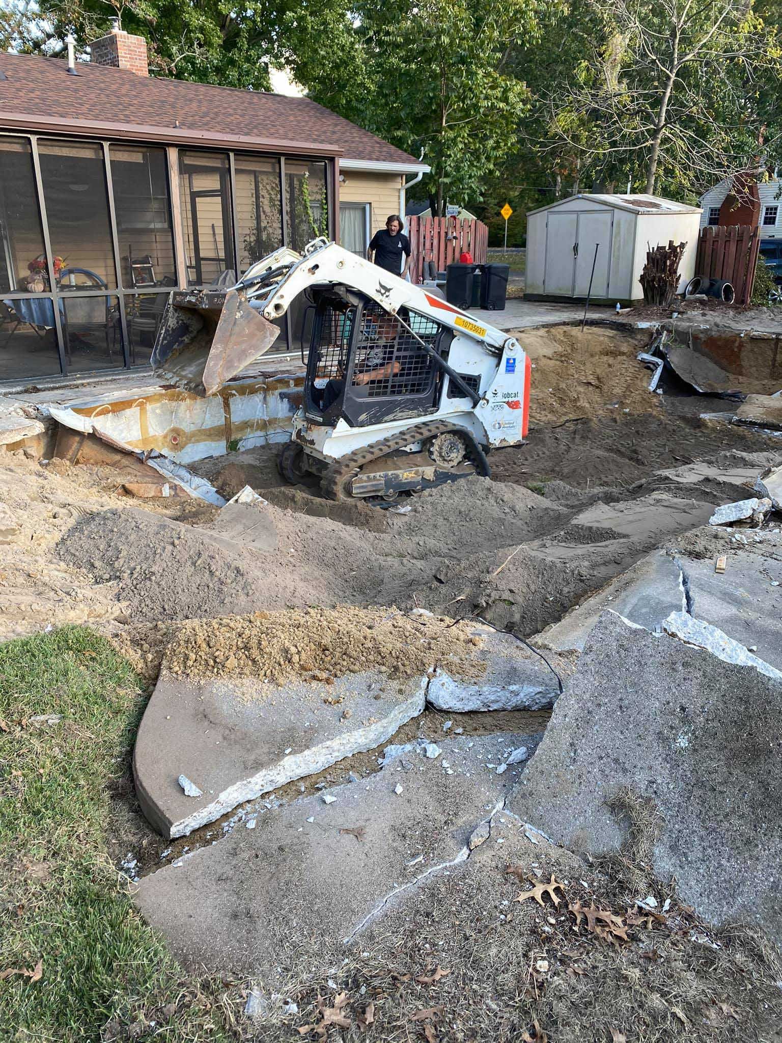 Bobcat skid-steer in a construction site; removing concrete next to a house. Soil and debris surround it.