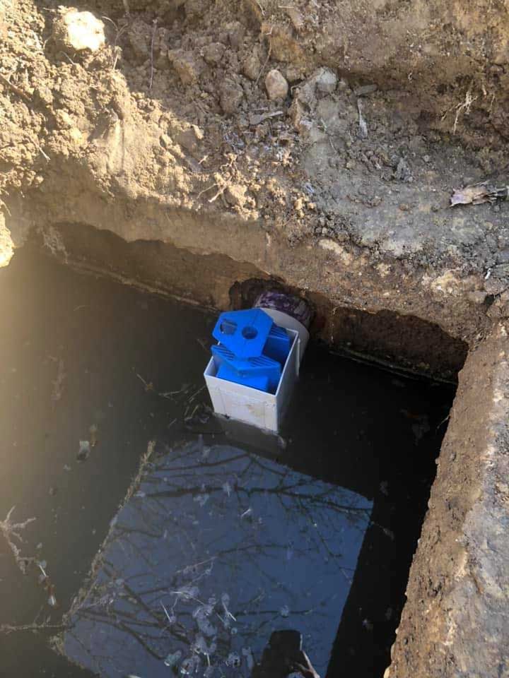 A blue pump inside a white box in a hole filled with water. Dirt surrounds the hole.