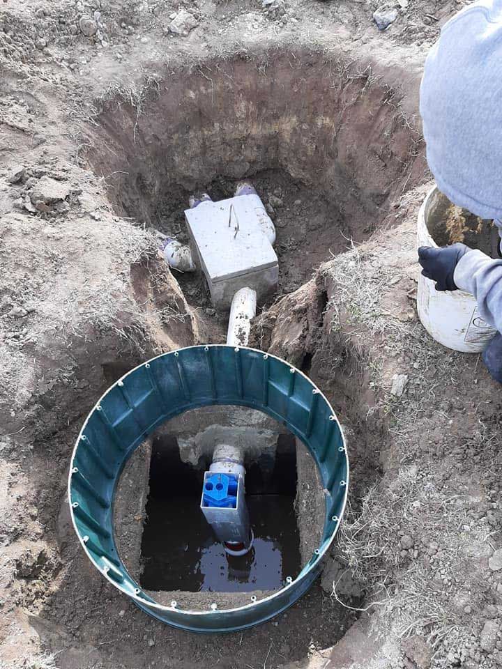 Person in gray jacket near a partially buried septic system, with green ring, pipes, and concrete blocks.