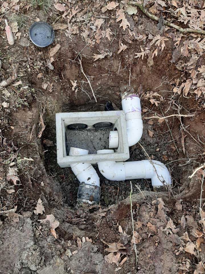 White plumbing pipes and a box-shaped structure in a hole in the ground, surrounded by dirt and fallen leaves.