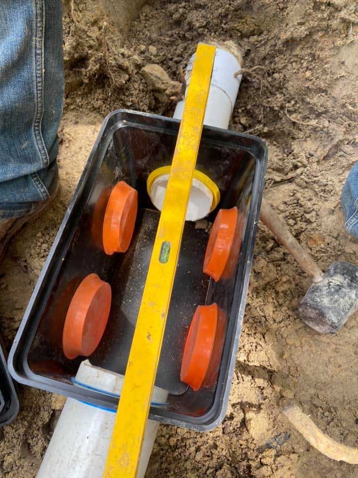 Level checking plumbing installation in dirt. Orange caps, yellow level, white and gray pipes.