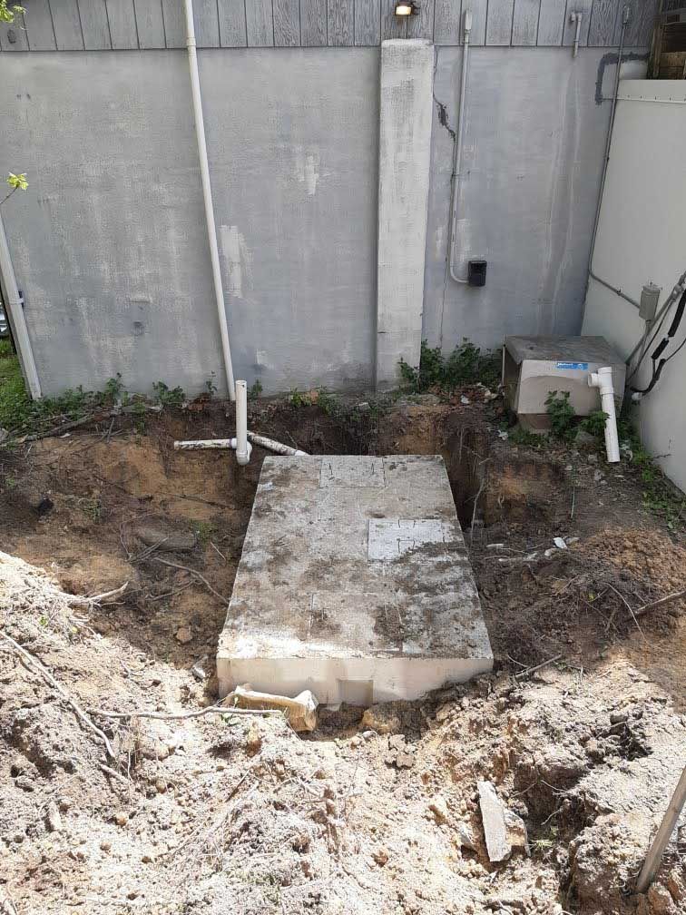 Concrete structure in a dug-out area, against a gray wall. White pipe and electrical box visible. Dirt surrounds it.