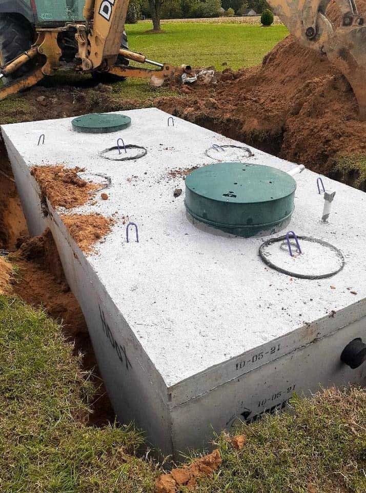 Concrete septic tank in a partially dug trench, with a backhoe in the background.