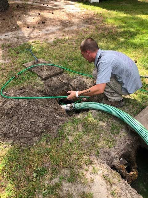 Man working near an open hole in the ground, with a green hose, in a grassy area.