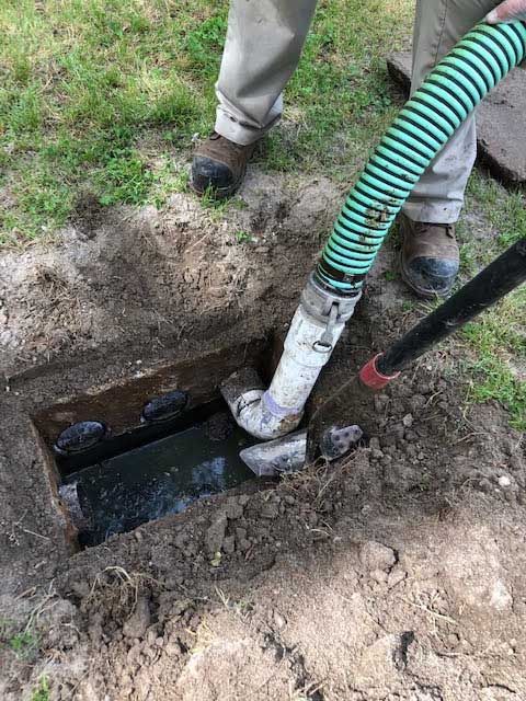 Person pumping septic tank with a hose in a dirt-dug hole; green grass background.