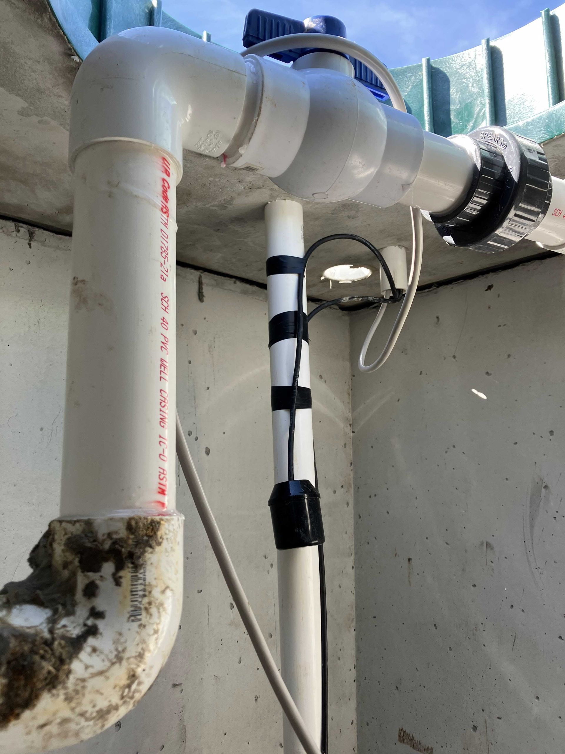White PVC pipes with a blue valve and black sensor, in a concrete enclosure.