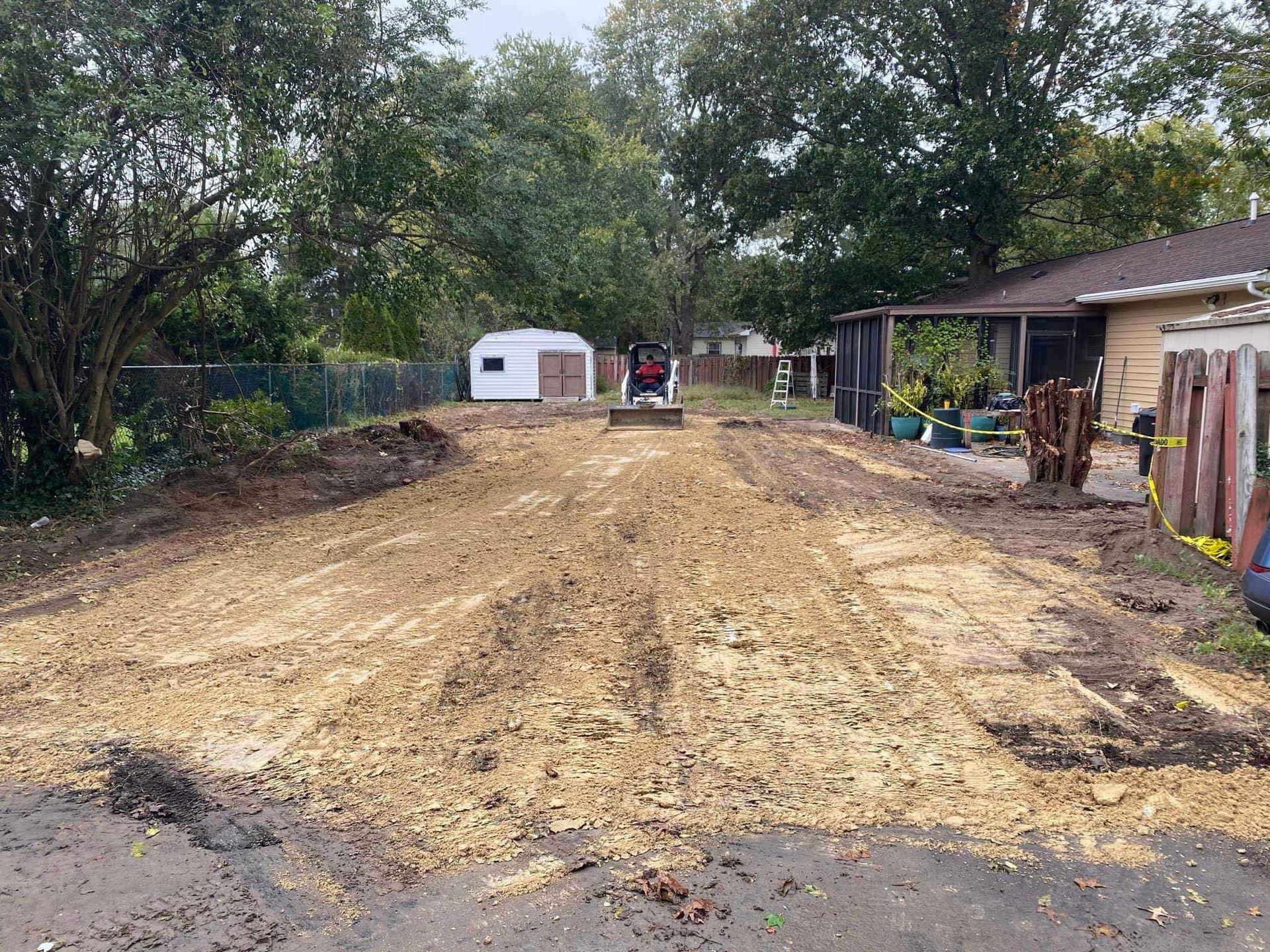 Dirt-covered yard with a small building in the back. A small excavator is in the middle of the yard.