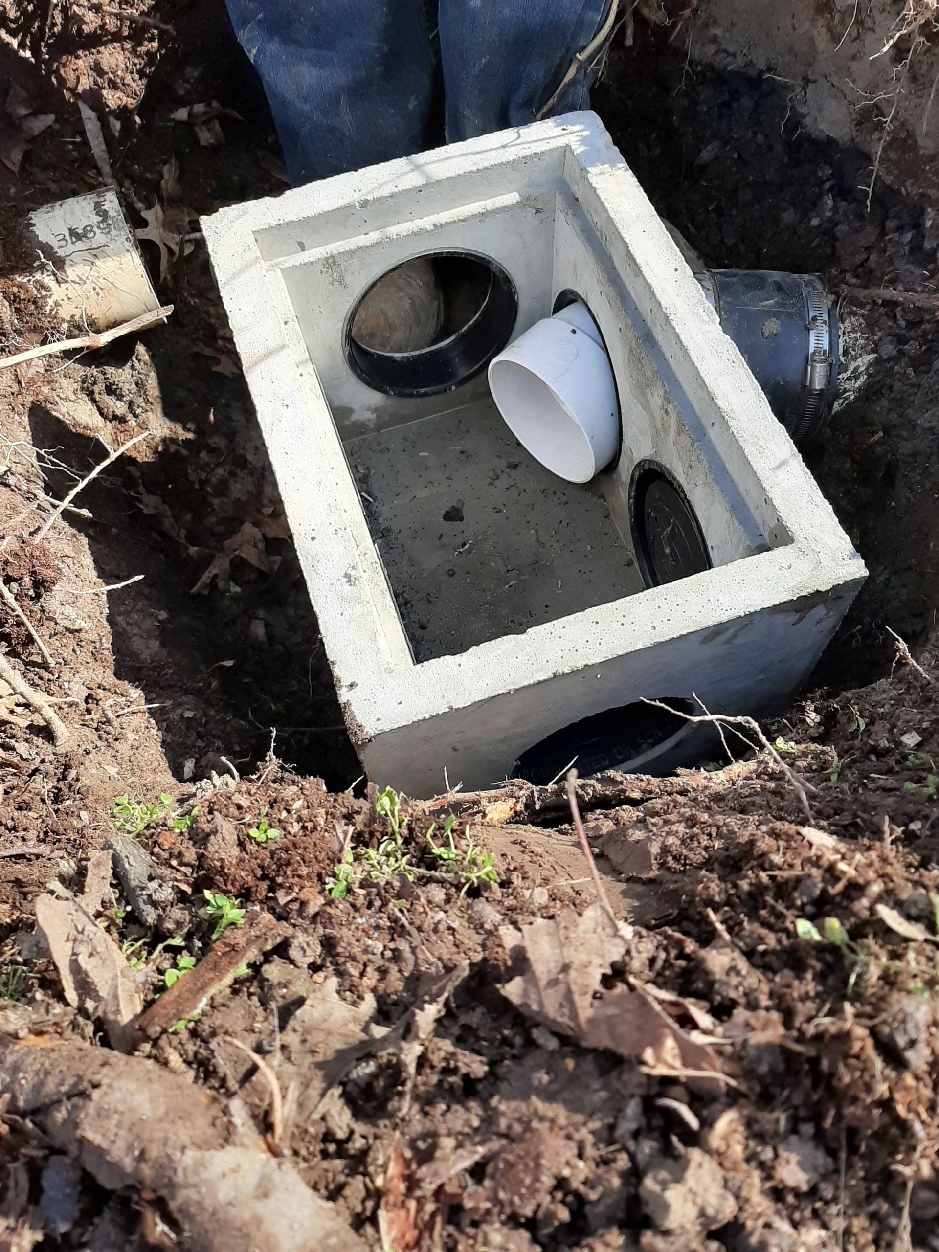 Concrete drainage box with pipes in a dirt trench.