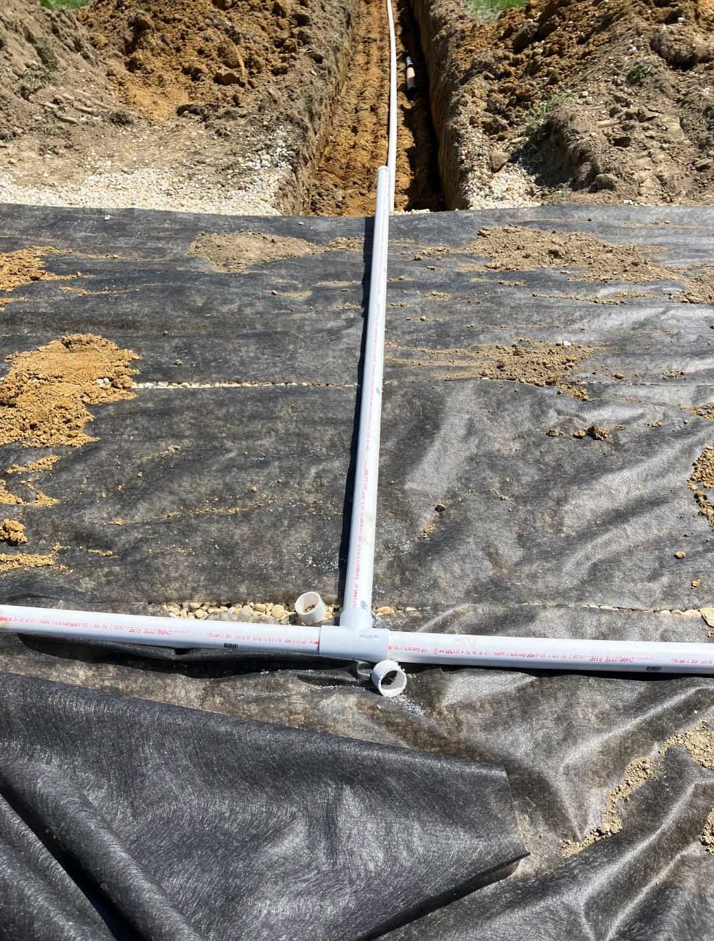 A white PVC pipe in a trench, connected with a T-fitting, on black fabric. Dirt in background.