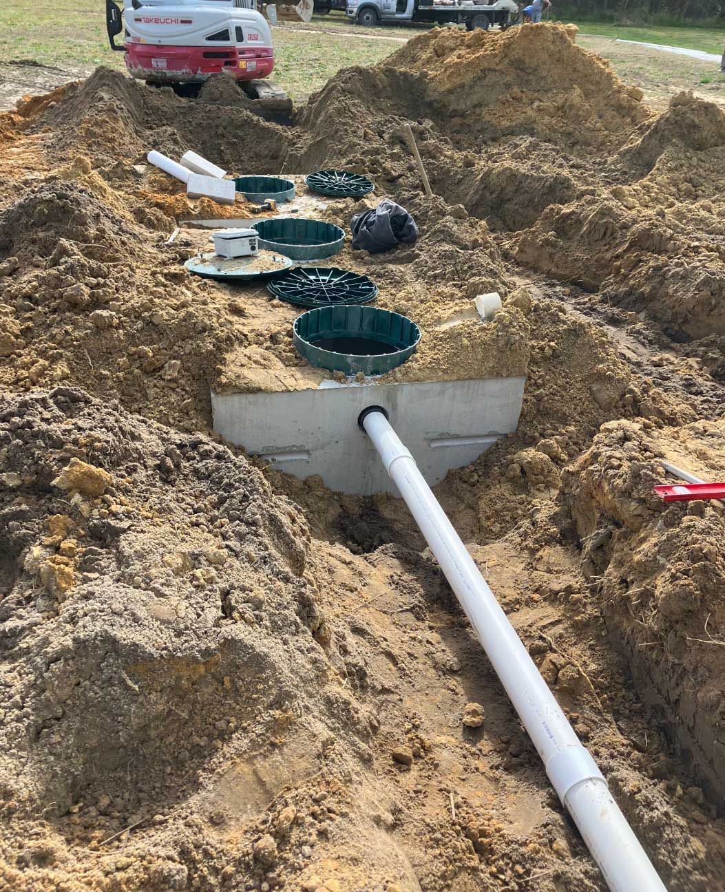 Septic system installation in progress; pipes and tanks exposed in a trench, surrounded by soil.