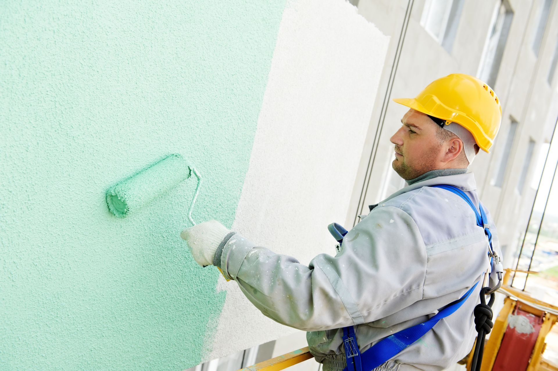 Worker in yellow hard hat painting a wall with a green roller indoors
