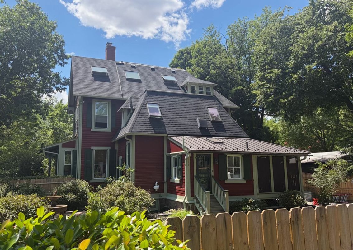 A red Victorian house with a steep roof and porch, surrounded by trees and a wooden fence.