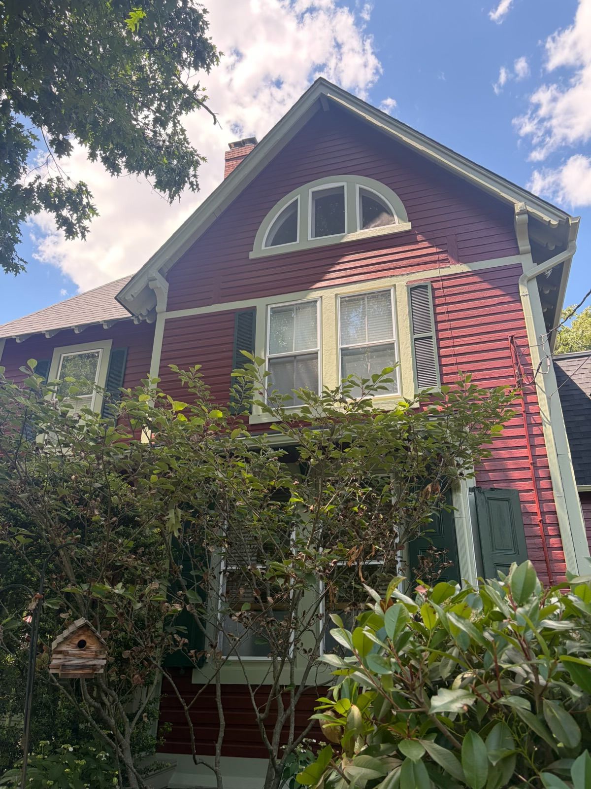 Victorian-style red house with white trim, arched attic window, and lush green shrubs in front