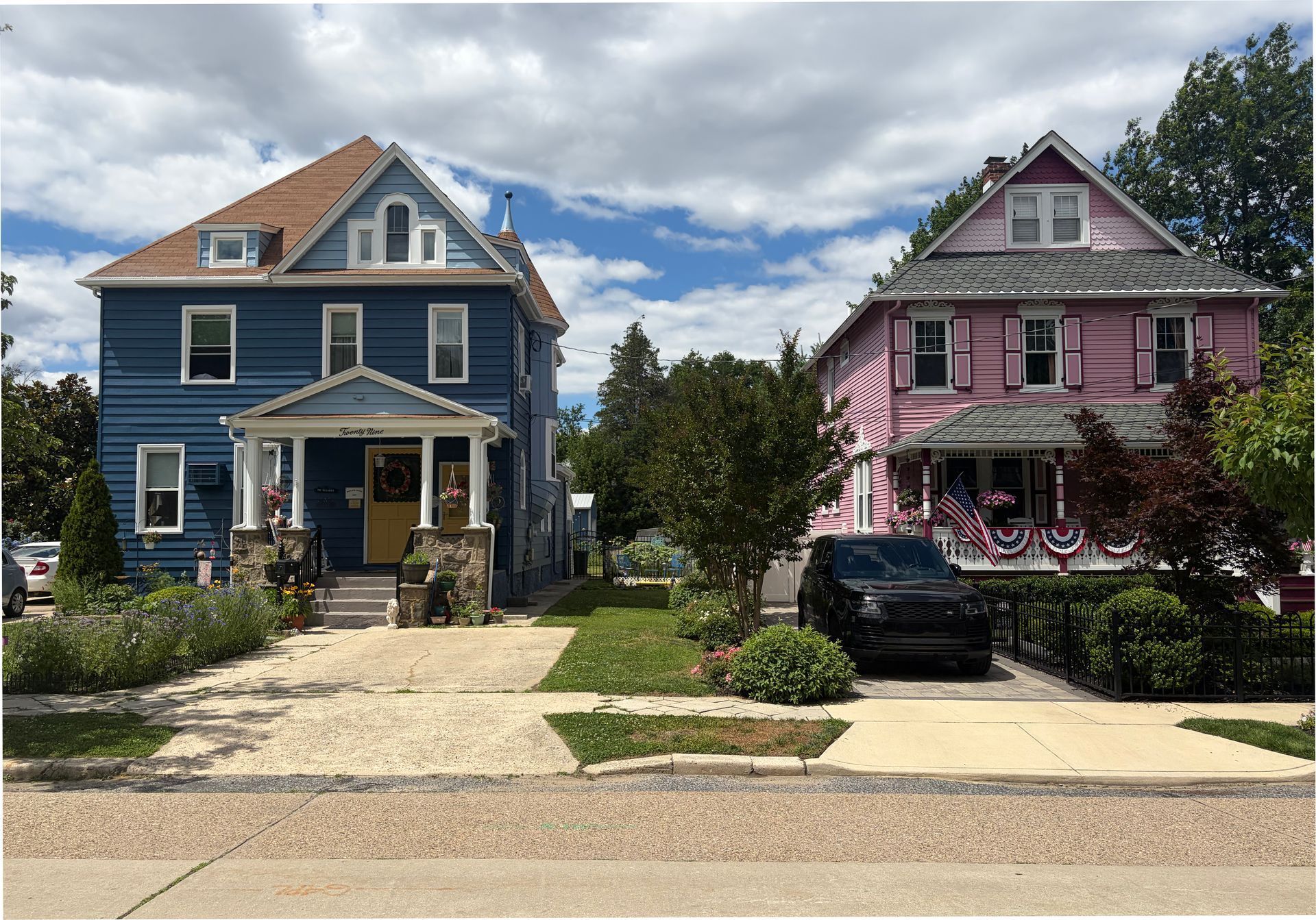 Two colorful Victorian houses, one blue and one pink, beside a sidewalk under a partly cloudy sky.