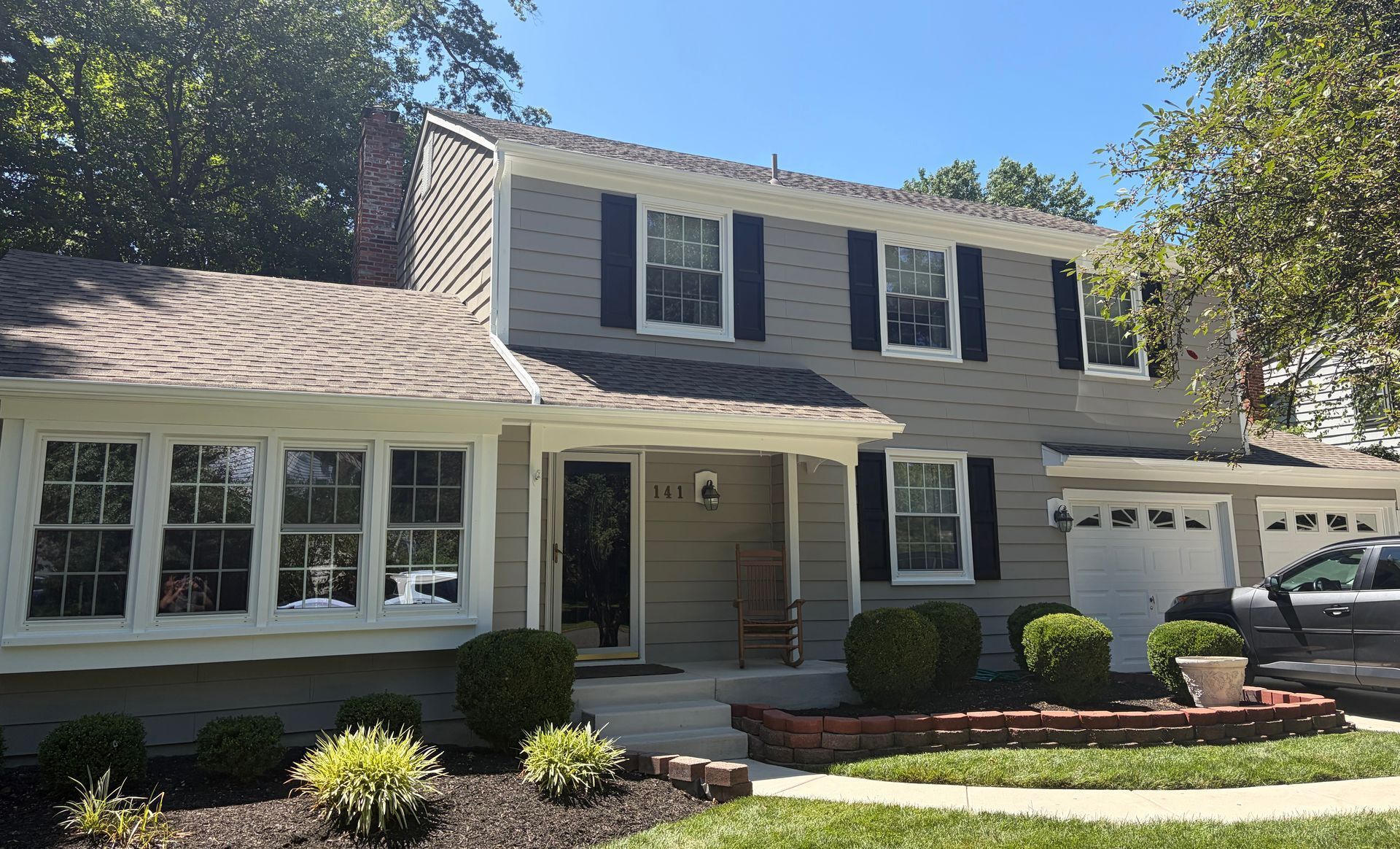 Two-story suburban house with white siding, dark shutters, and a front porch under a sunny sky