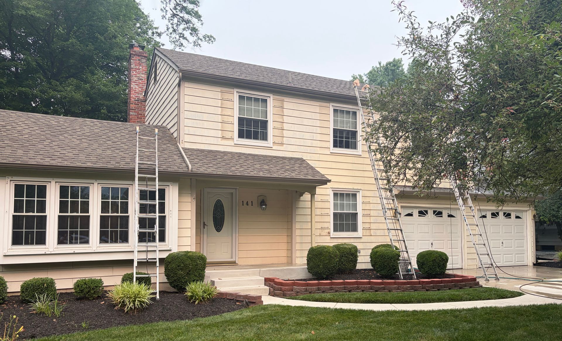 Two-story beige house with white trim, a front porch, attached garage, and manicured shrubs