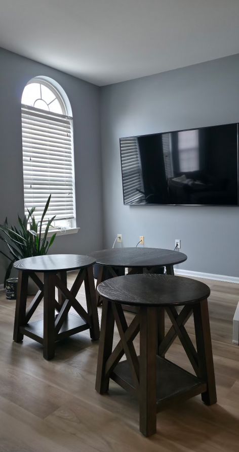 Small dining nook with round black table, two stools, wall-mounted TV, and arched window in gray room