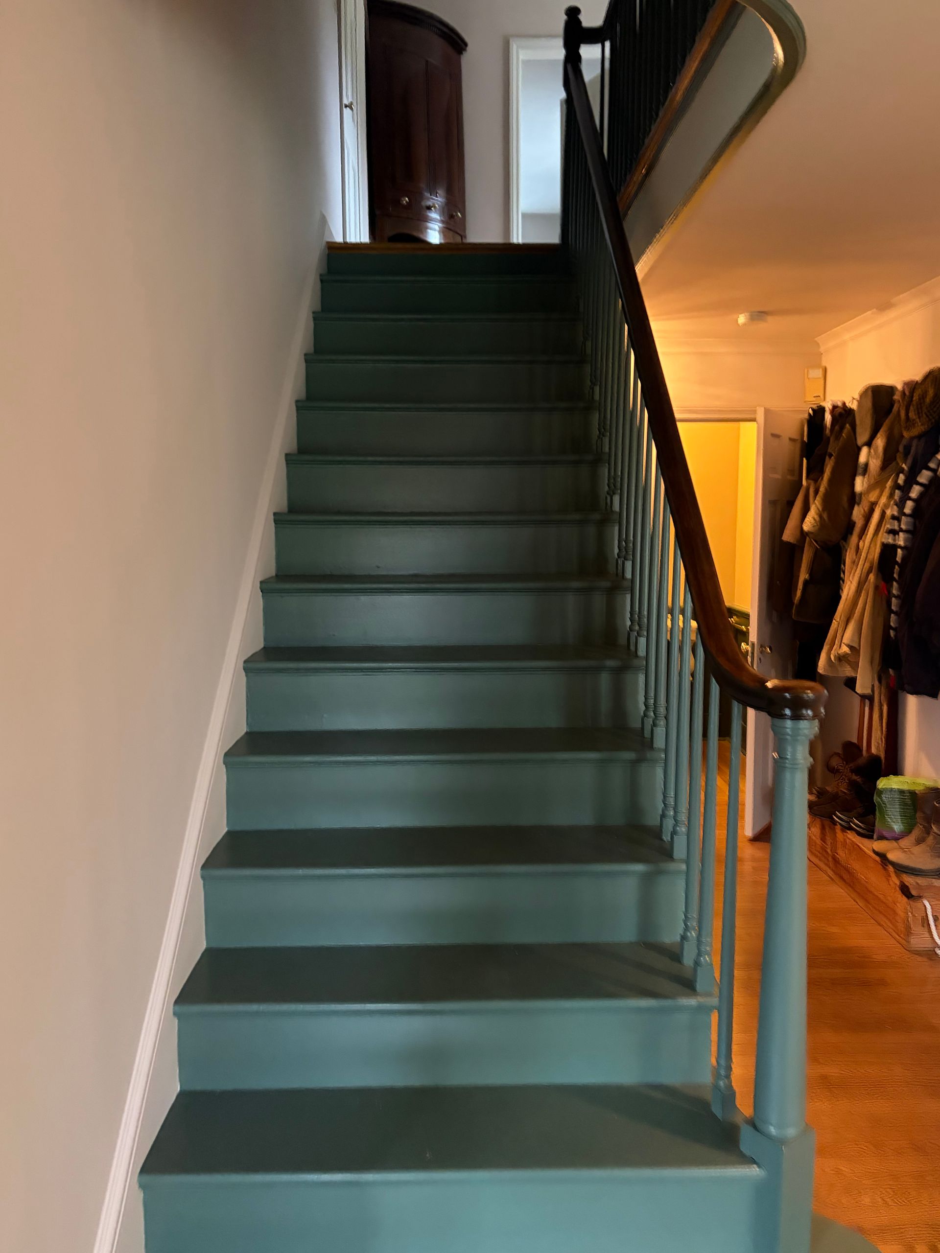 Green staircase in a narrow hallway beside a warm-lit room with coats hanging on the right.