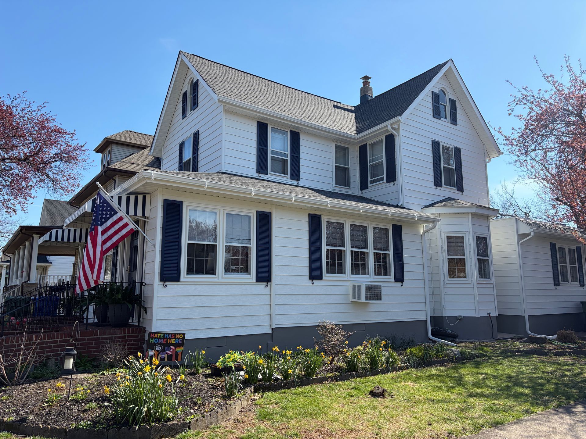 White two-story house with black shutters, an American flag, and a front garden on a sunny day