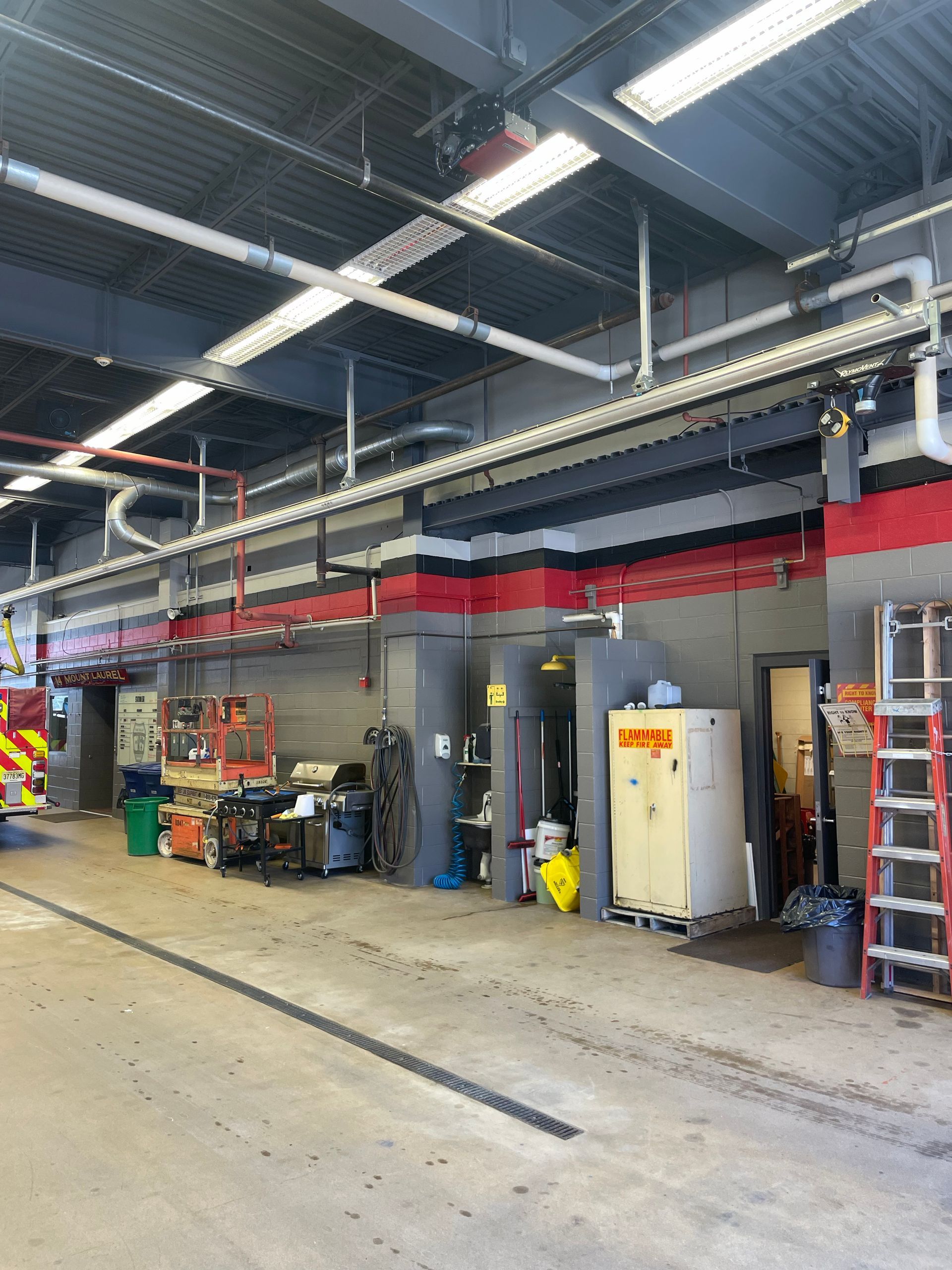Industrial garage with open bays, red and gray walls, tools, ladders, and overhead piping under bright lights