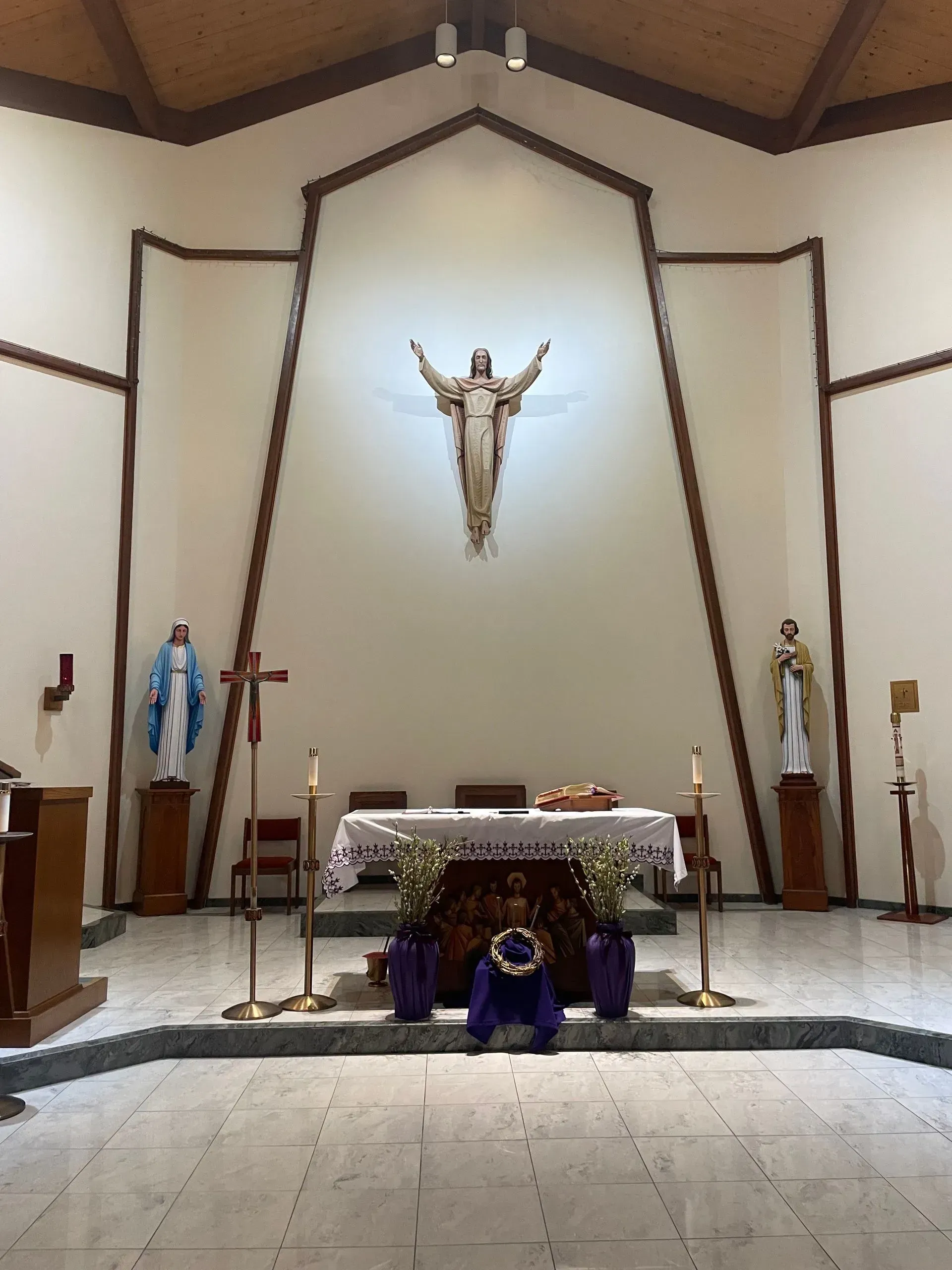Interior of a church; altar with Jesus sculpture, statues, and floral decorations. Beige walls, wooden trim, and grey floor.