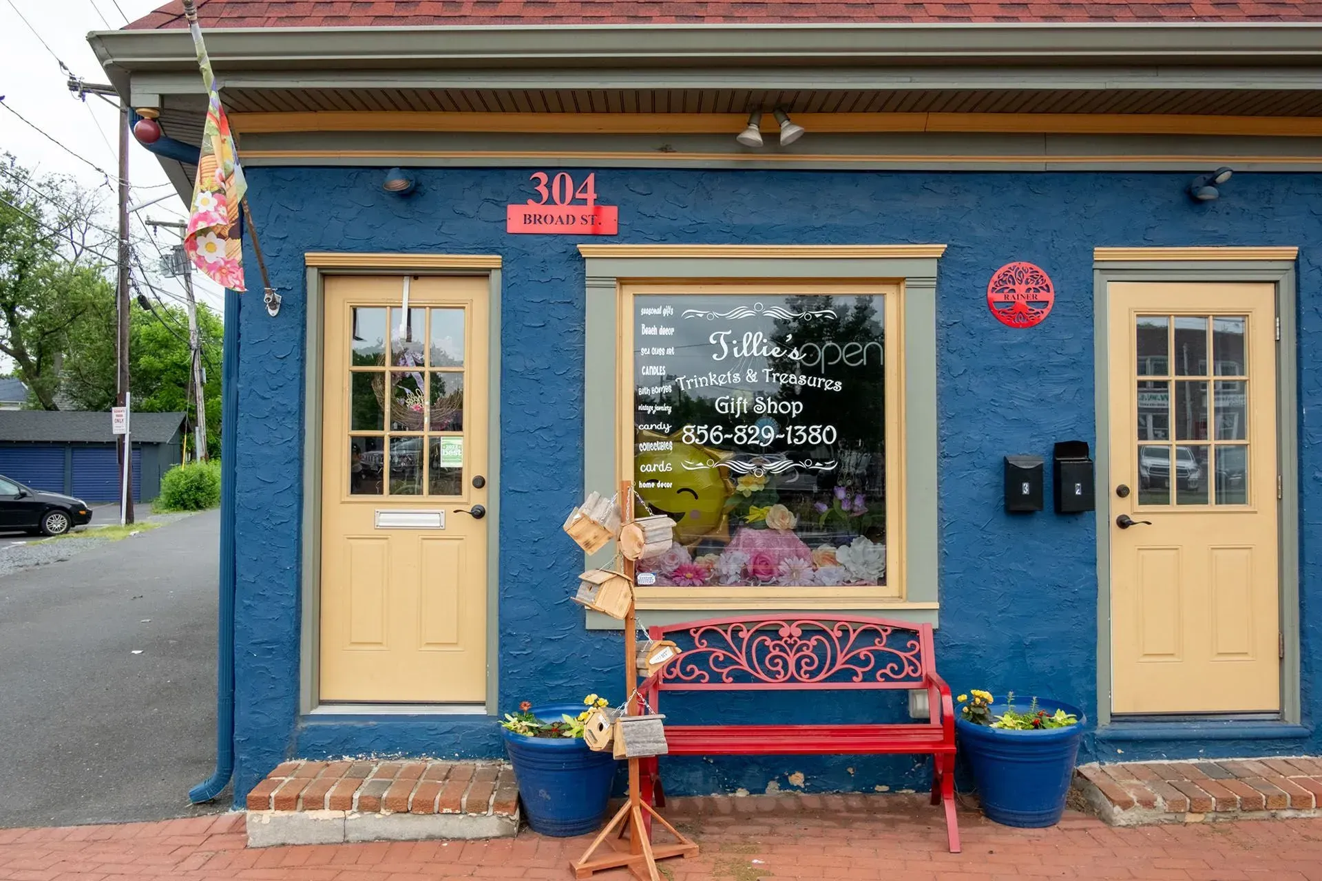 Blue storefront with yellow doors, bench, flower pots, and a window display on a brick sidewalk.