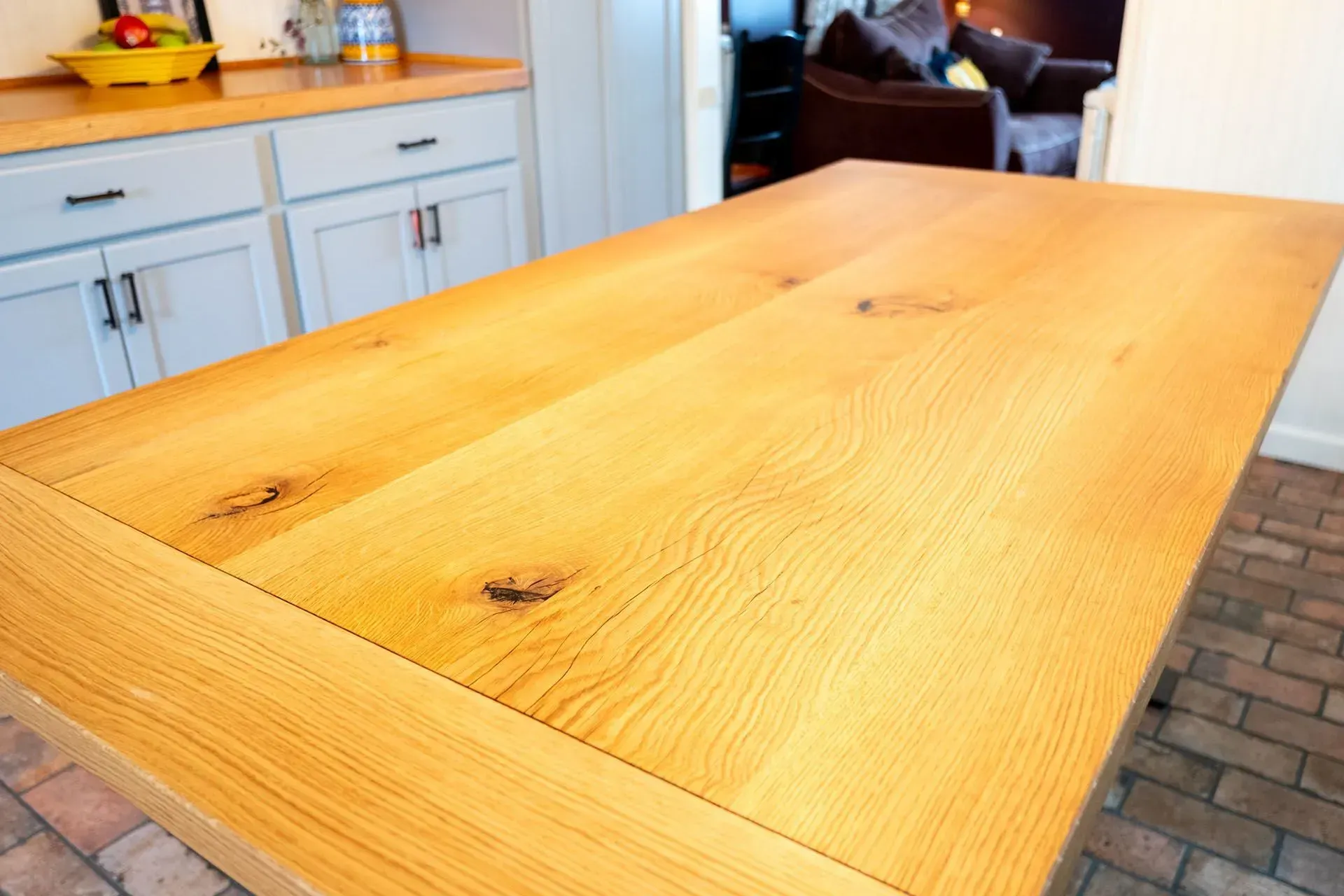 Close-up of a large, light-colored wooden kitchen island. Cabinets and brick flooring are visible.