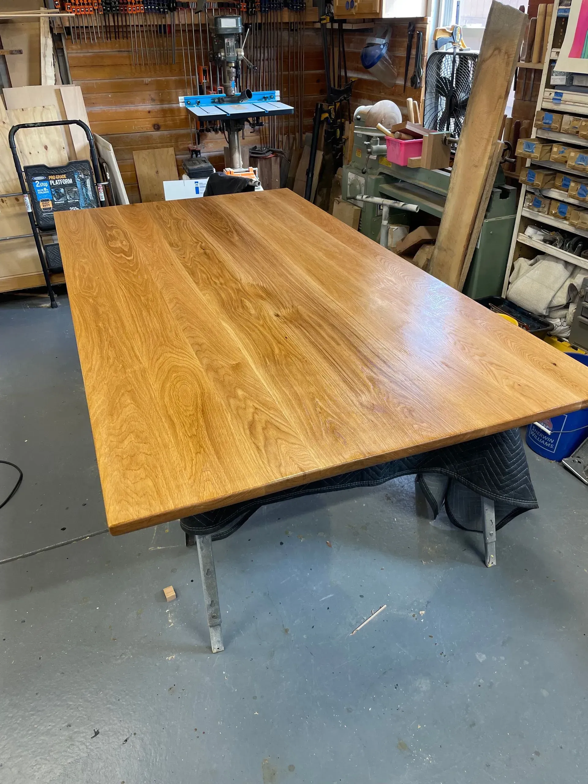 Large, stained wooden tabletop set on a dark support in a workshop, with tools and equipment in the background.