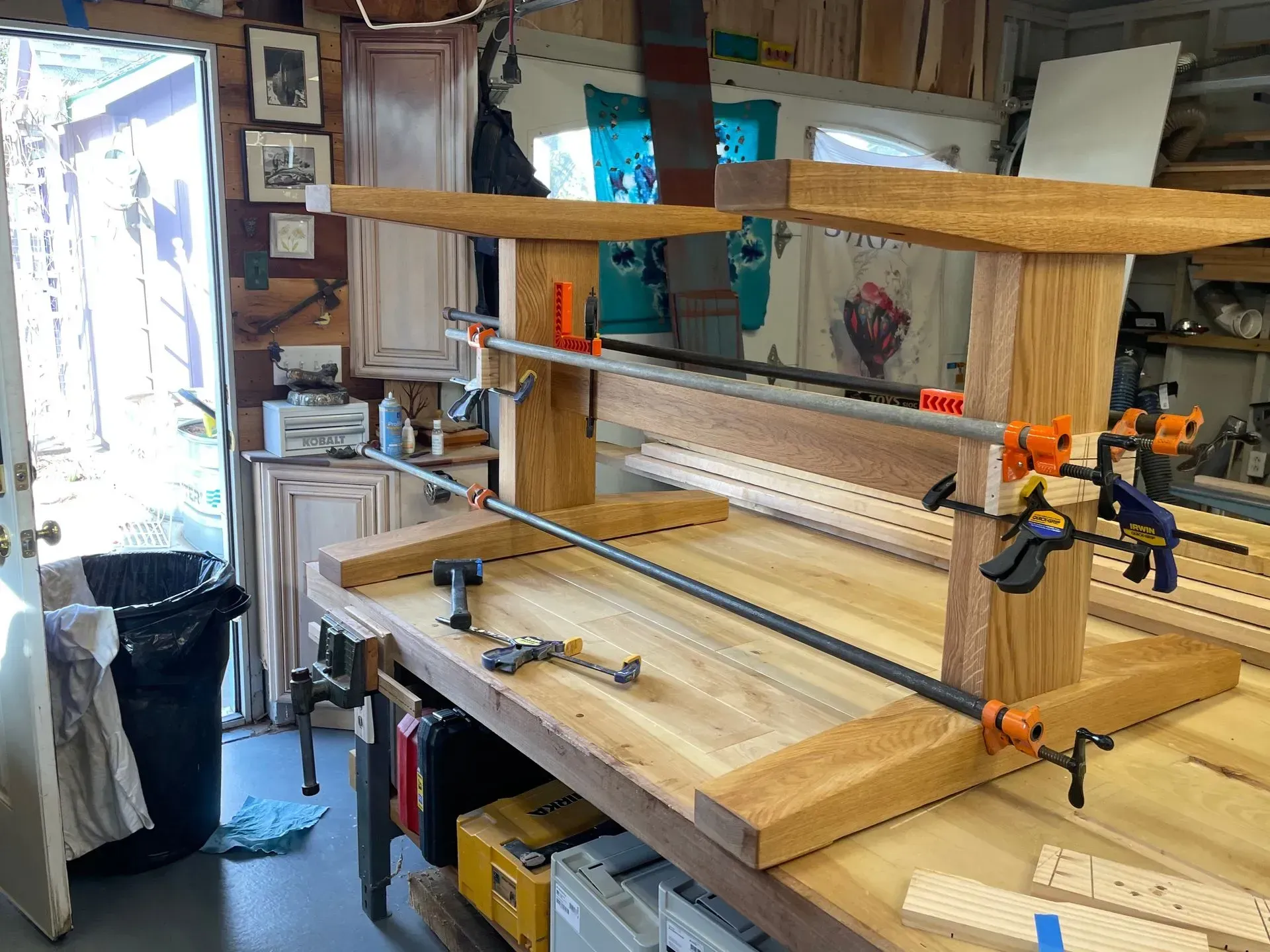 A wooden table being assembled with clamps in a workshop. Light wood tones, tools, and a cluttered workspace.