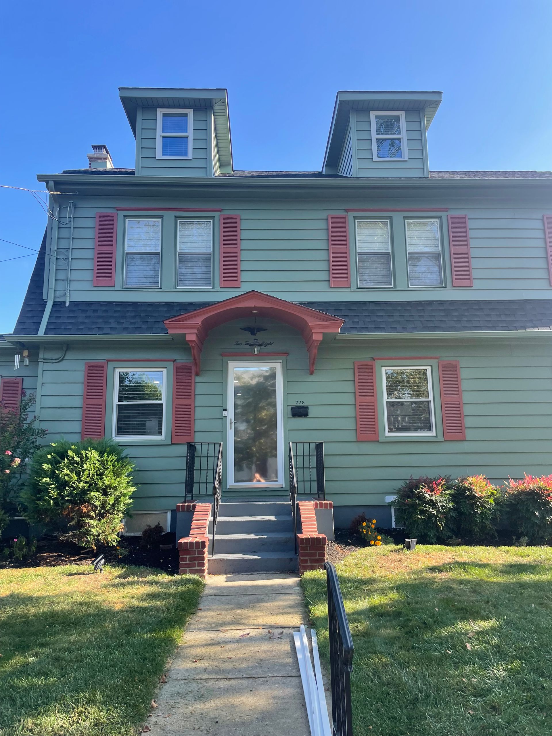 Green two-story house with red shutters and dormers. Front steps lead to a glass door. Clear blue sky.