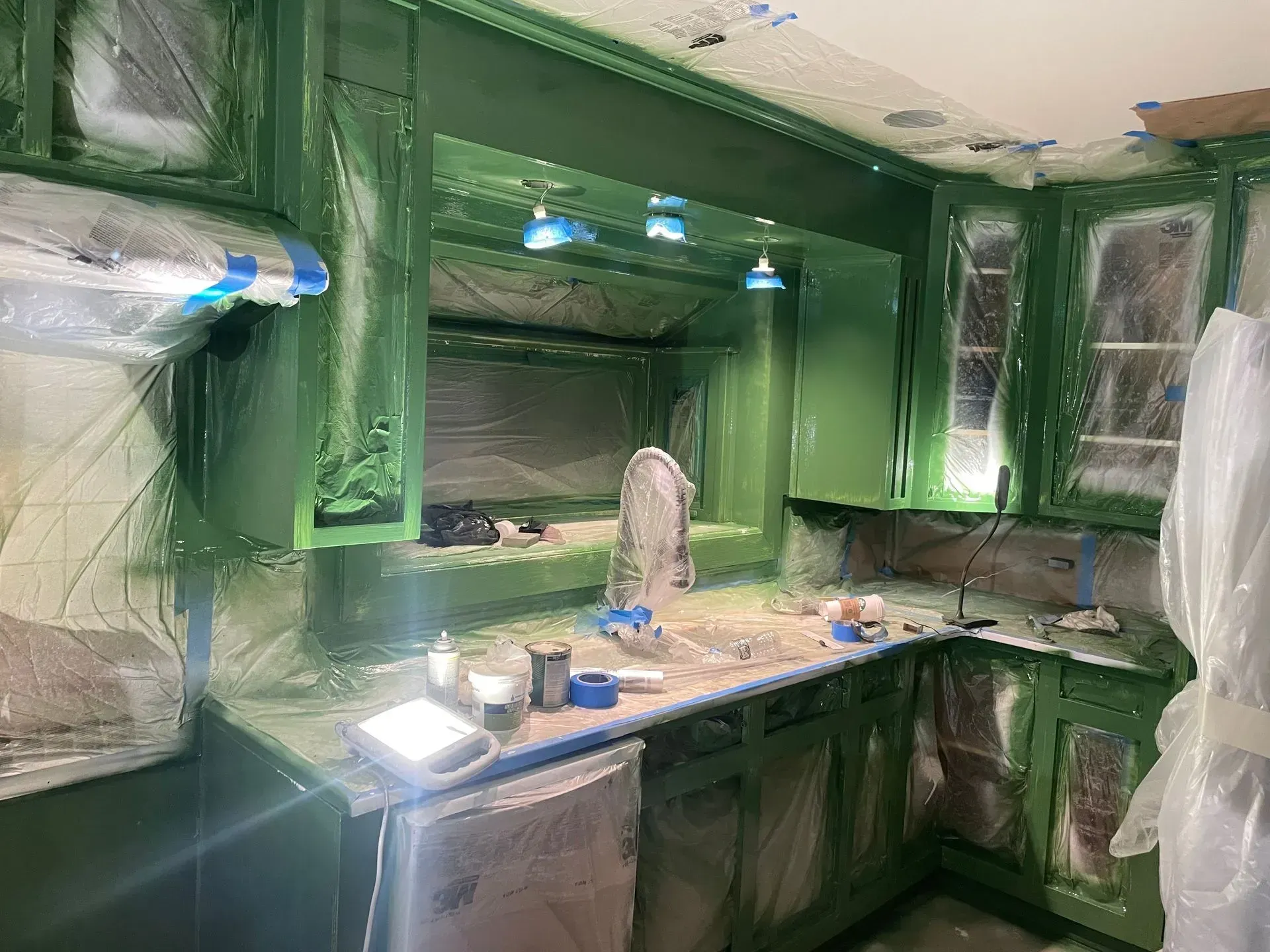 Kitchen being painted green, covered in plastic, with sink, cabinets, and lights visible.